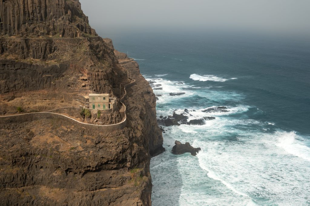 Clifftop winding path along rugged dark rock, a small concrete building perched near the edge above churning ocean.