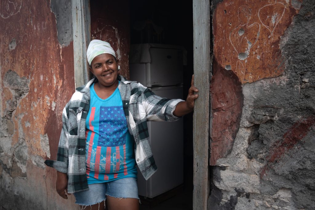 Smiling woman in a blue shirt with an American flag design and a plaid shirt, standing in a doorway of a worn, red-brick building.
