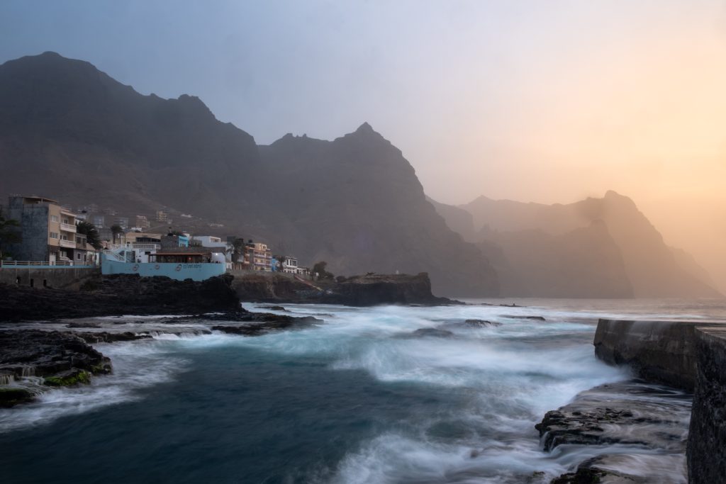 Coastal town at the base of dark mountainous cliffs during sunset; waves crash against rocks.