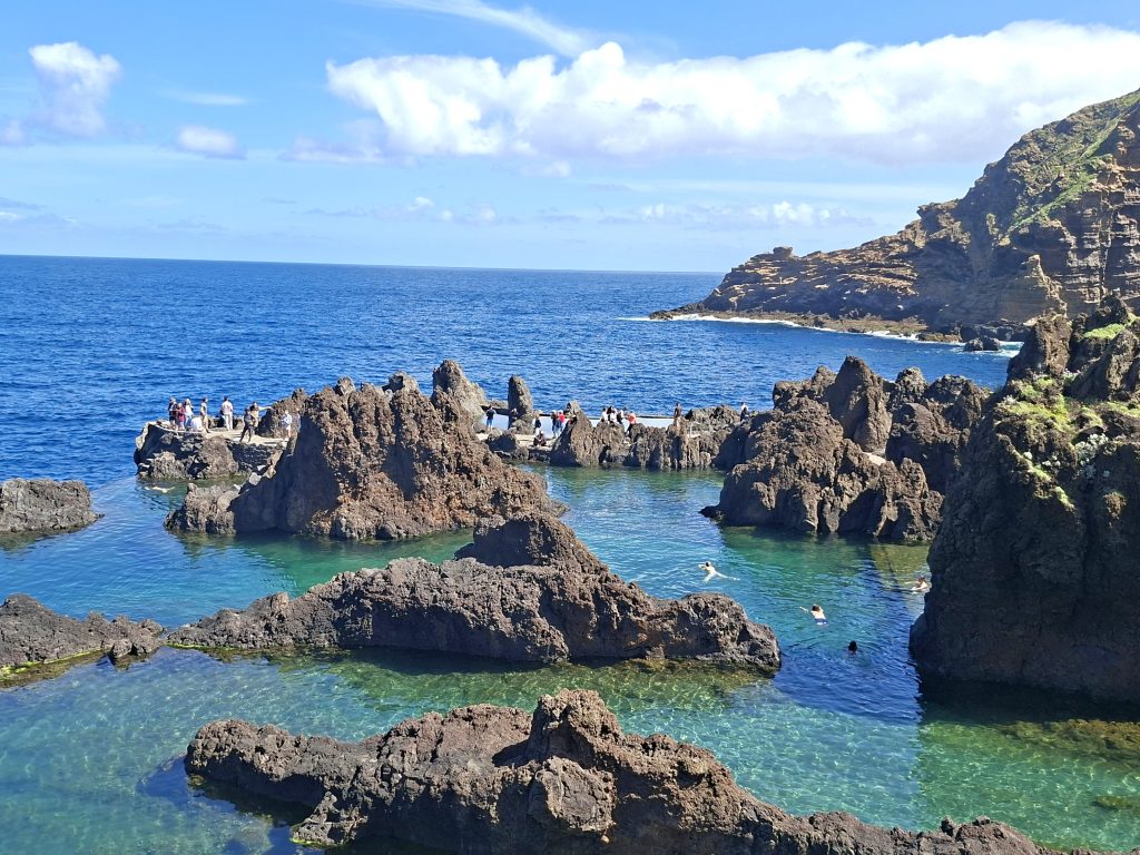 Group of people exploring jagged lava rocks along a bright blue sea under a clear sky, with rocky cliffs to the right.