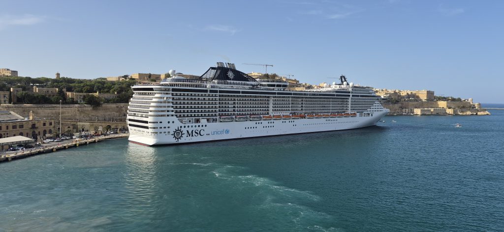 Large white MSC cruise ship docked near a historic fortress town, blue sea and clear sky behind it, UNICEF branding visible on hull