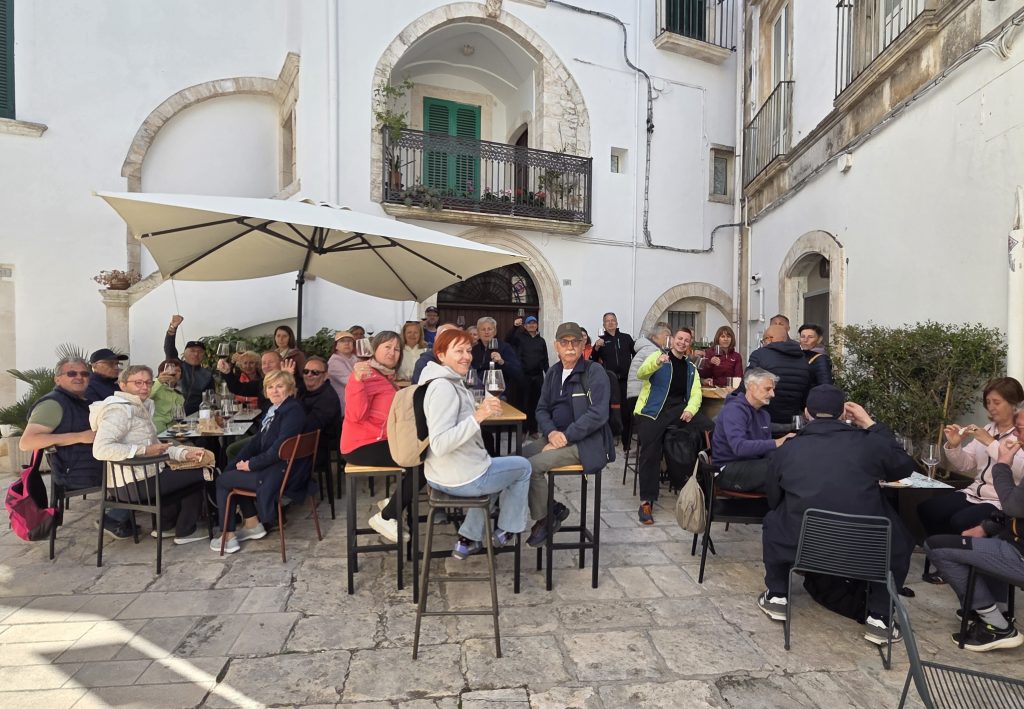Group of people at an outdoor cafe raising glasses in a sunny town square with white buildings and arches behind them.