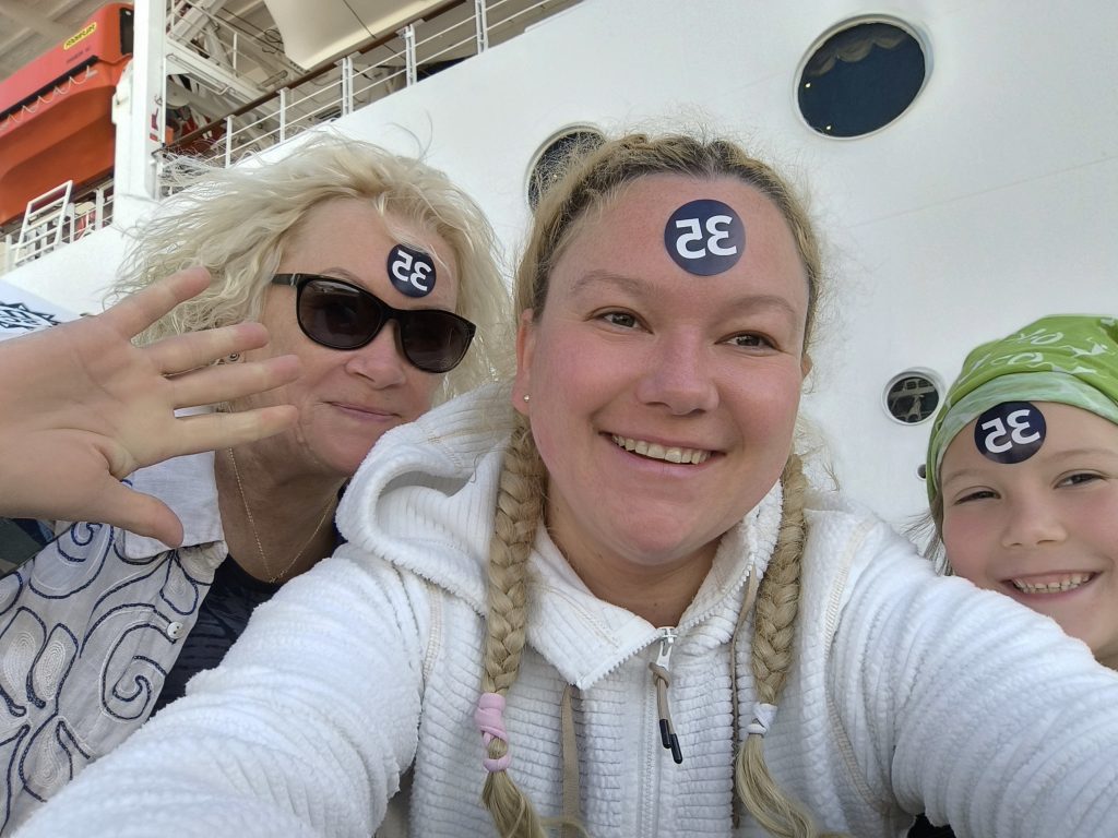 Three people taking a selfie on a ship deck, with blue circular '35' stickers on their foreheads behind sunglasses and braids visible in the foreground.