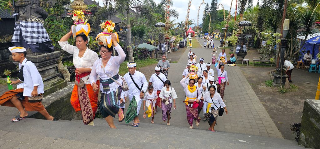 Group of Balinese people in white clothing walking up steps, balancing decorative offerings on their heads during a temple festival.