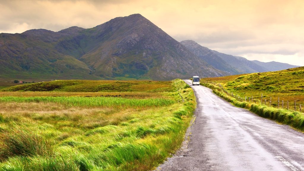 Narrow rural road winds toward green fields and towering mountains, a white bus in the distance.