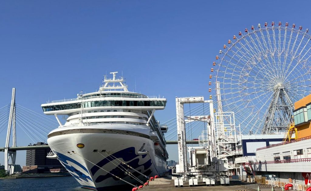 Large white cruise ship docked at a port with a Ferris wheel and blue sky behind.