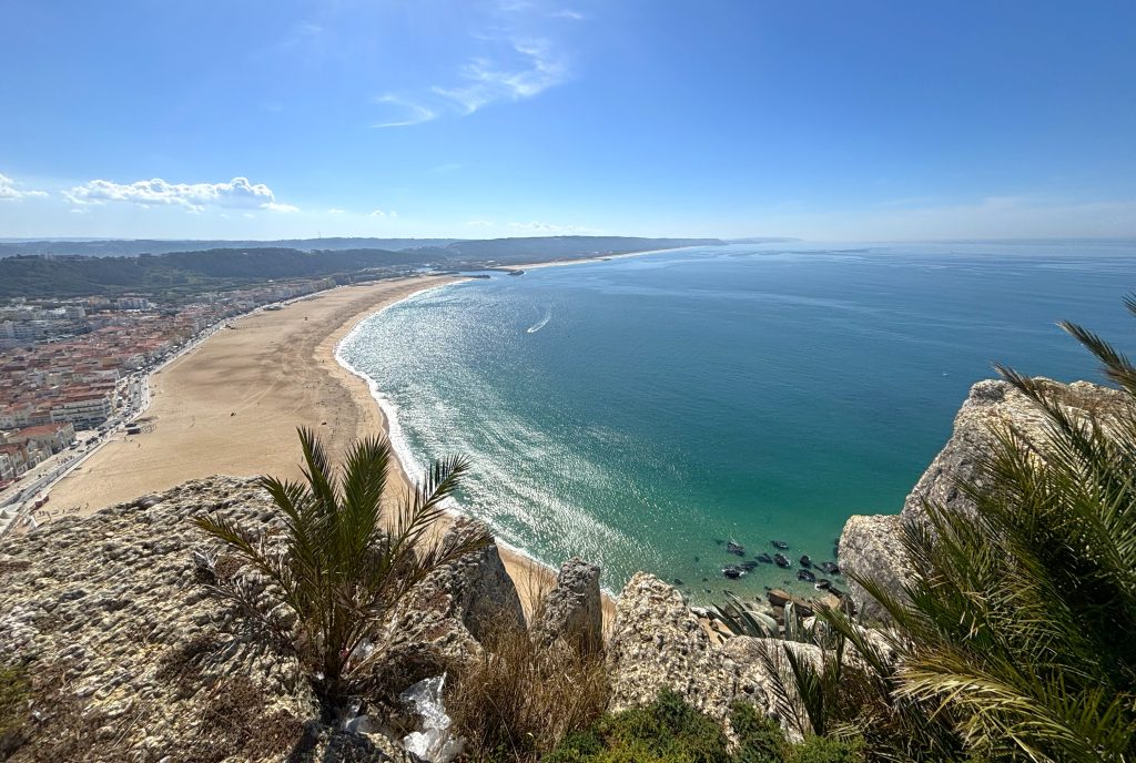 A coastal town along a curved sandy beach, seen from a rocky cliff with blue sea and clear sky above.