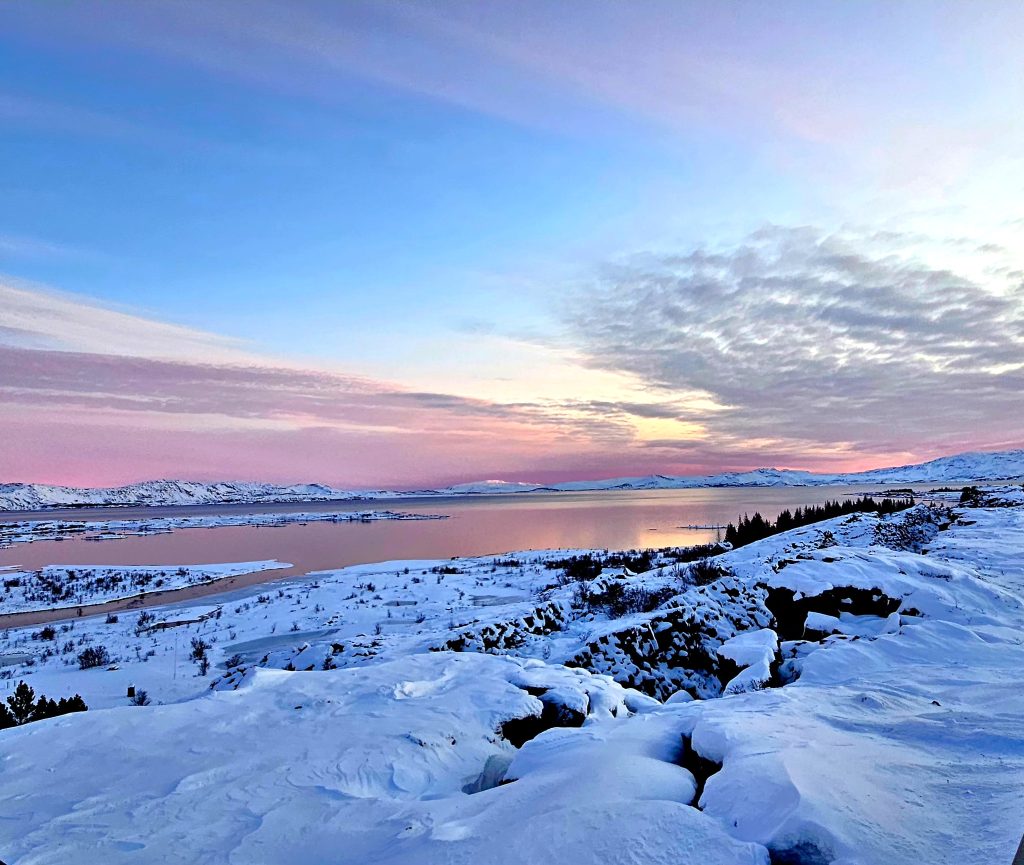 Snow-covered hillside beside a calm lake under a pastel blue-pink sunset sky with distant mountains and sparse trees on the right.