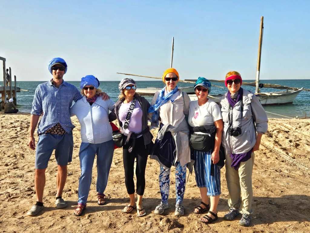 Six adults pose arm-in-arm on a sandy beach with a boat and calm sea behind them on a sunny day, wearing colorful headscarves and sunglasses.
