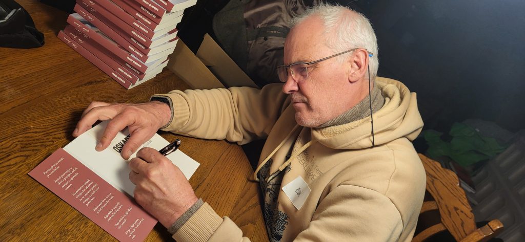 Senior man wearing a beige hoodie signs a book at a wooden table, with a stack of pinkish-red books nearby, during a book signing.