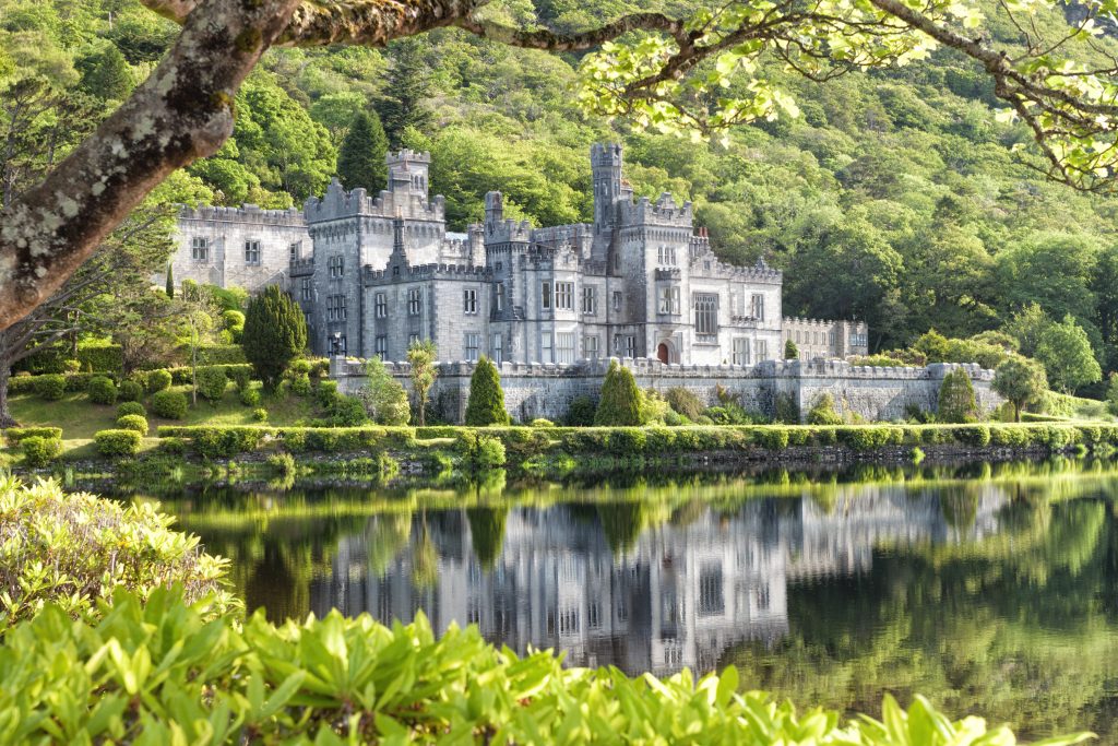 Castle-style stone mansion with crenellated towers sits beside a calm lake, surrounded by lush green trees.