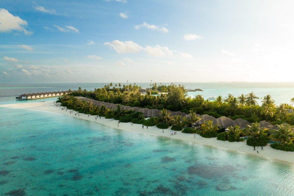 Aerial view of a tropical island resort with overwater bungalows, palm trees, and a white sand beach along turquoise water.