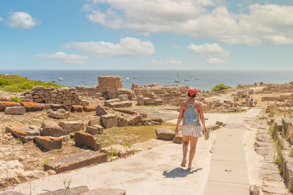 Tourist with a blue backpack walking along ancient stone ruins by the sea with boats on the horizon.