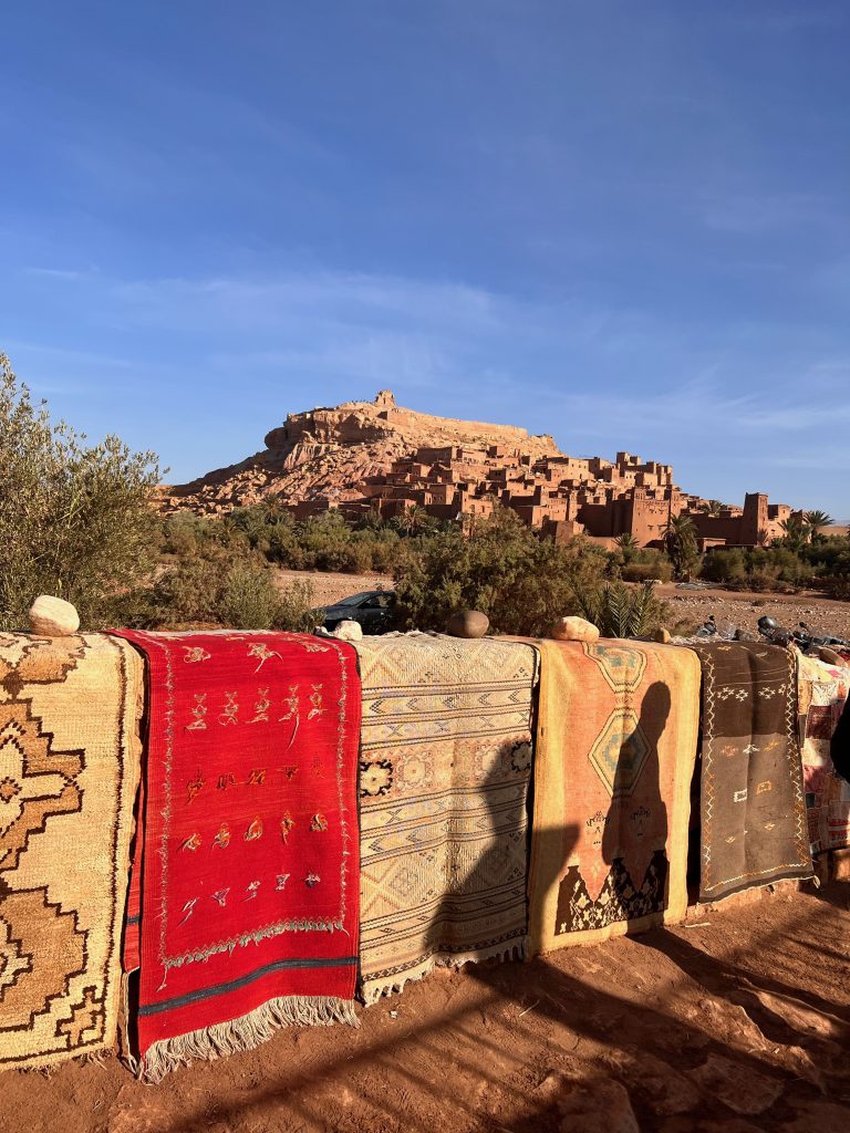 Colorful woven carpets hang along a low wall in a desert setting with a clay village on a hill in the distance under a clear blue sky, olive trees in foreground