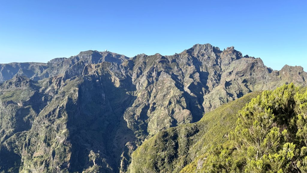 Rugged mountain range under a bright blue sky, with green shrubs in the foreground.