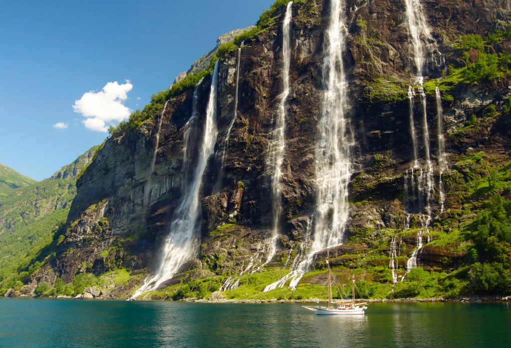 Tall waterfalls cascade down a rocky cliff into a blue-green bay, with a small boat near the shore under a clear sky.