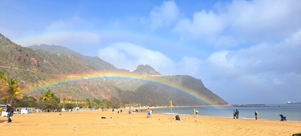 Beach scene with golden sand, palm trees, and a rainbow arching over rocky hills under a blue sky.