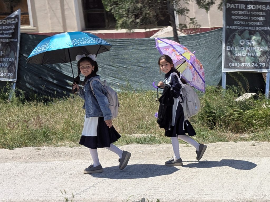 Two schoolgirls in uniform walking along a sunny street, each carrying a backpack and an umbrella.