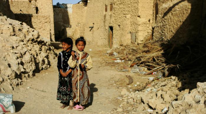 Majhni deklici v lokalnih nošah pozirata pred hišami iz blata. 