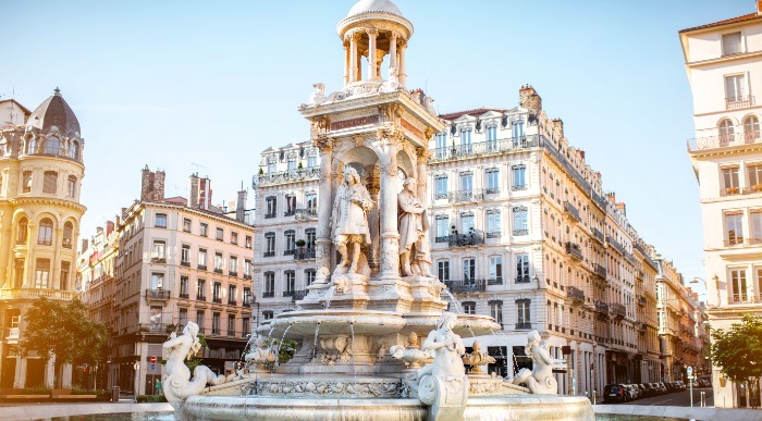 Sonce osvetljuje okrašen vodnjak na trgu Jacobins med zgodovinskimi stavbami v središču Lyona