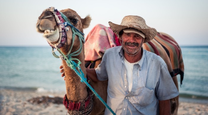 Gospod z slamnatim klobukom nasmejan stoji na plaži s kamelo ob rami. 