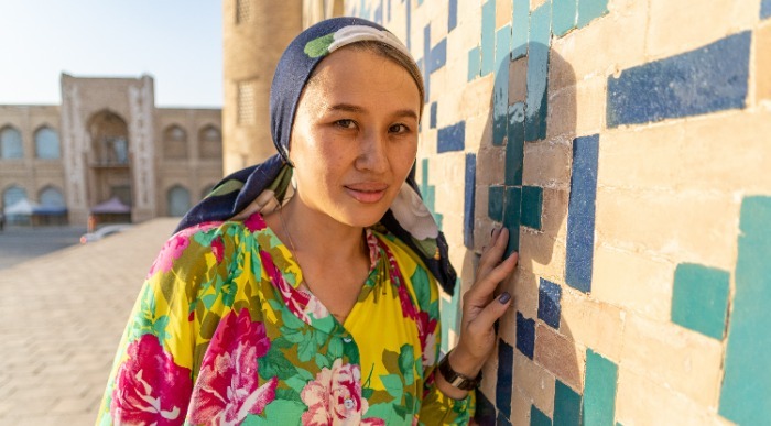 Ženska v barviti tradicionalni obleki stoji ob starodavno okrašenem zidu s keramičnimi vzorci  v Uzbekistanu