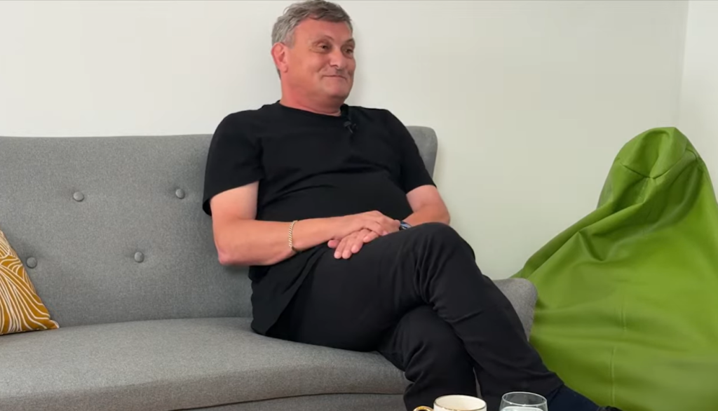 Man in a black T-shirt seated on a gray sofa with legs crossed and hands folded; a green bean bag sits to his right.